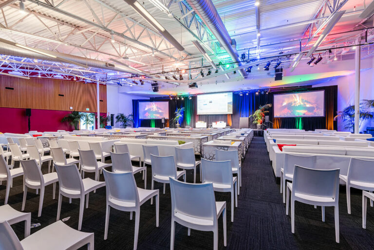 Rows of white chairs and lounge seating facing a stage and three screens in a museum event hall.