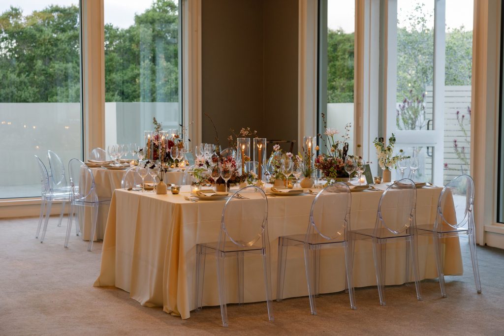 Reception table in cream linen with clear ghost chairs, taper candles in glass hurricanes, and autumn wildflower centerpieces beside tall windows