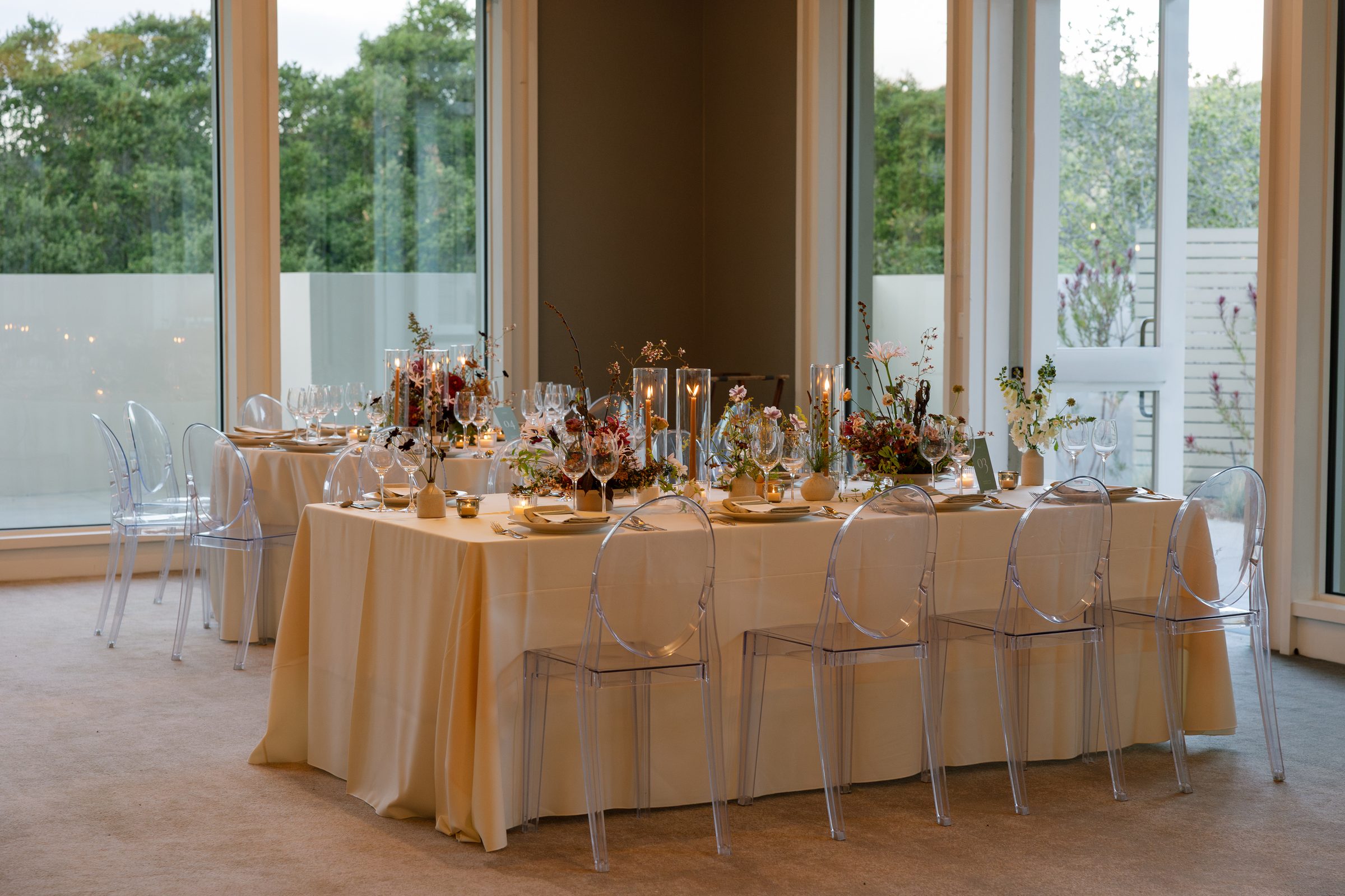 Reception table in cream linen with clear ghost chairs, taper candles in glass hurricanes, and autumn wildflower centerpieces beside tall windows